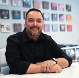 A man with dark hair and a beard, wearing a black shirt, sits at a desk and smiles. Colorful music album covers are displayed on the wall behind him.